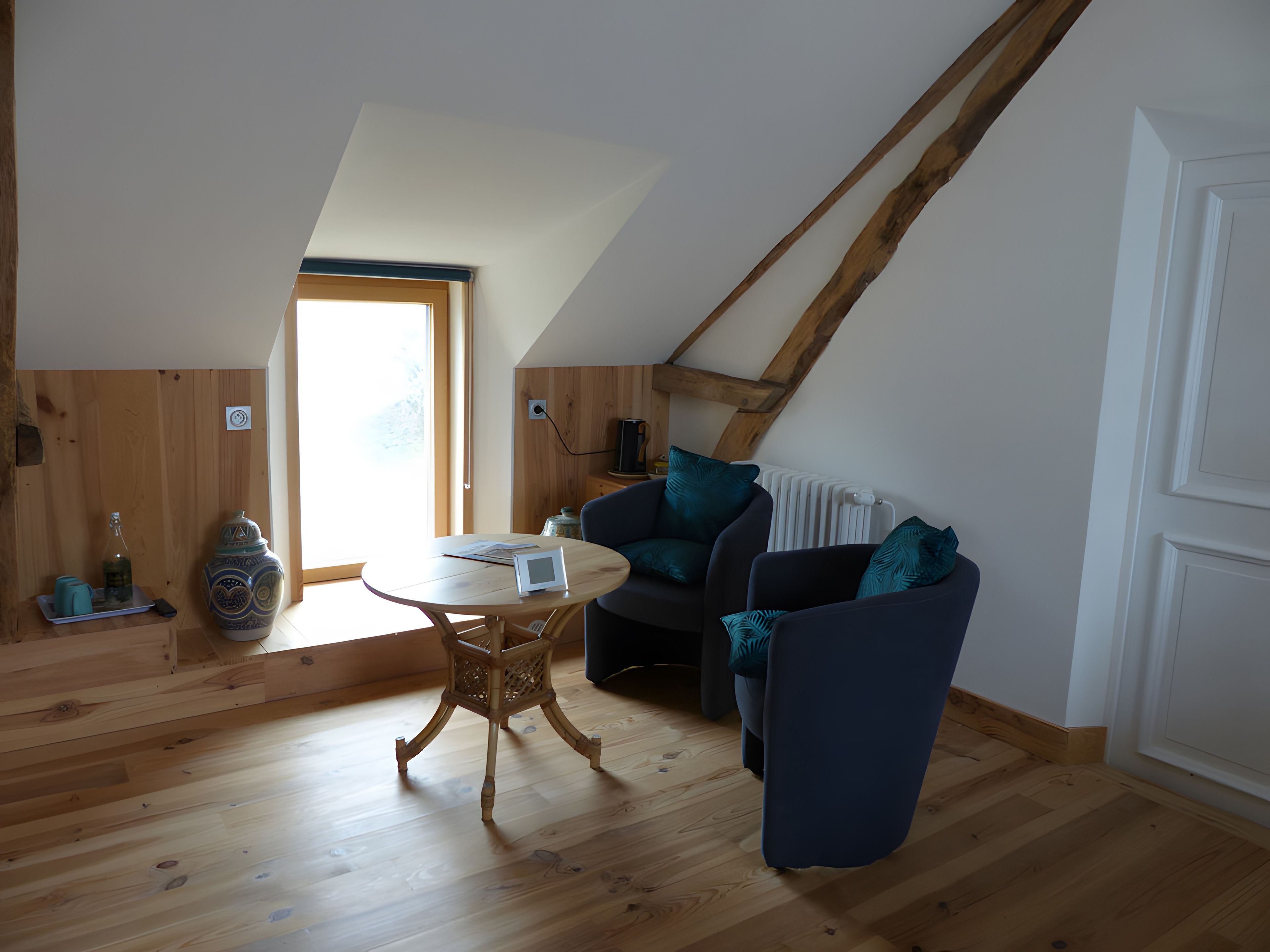 A cozy loft-style room with a sloped ceiling, wooden flooring, a round table, and a dark blue sofa with teal cushions.