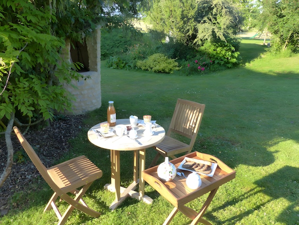 Una mesa redonda de madera con sillas, una botella, tazas y un plato con comida, en un espacio al aire libre con césped, árboles y un jardín de fondo.