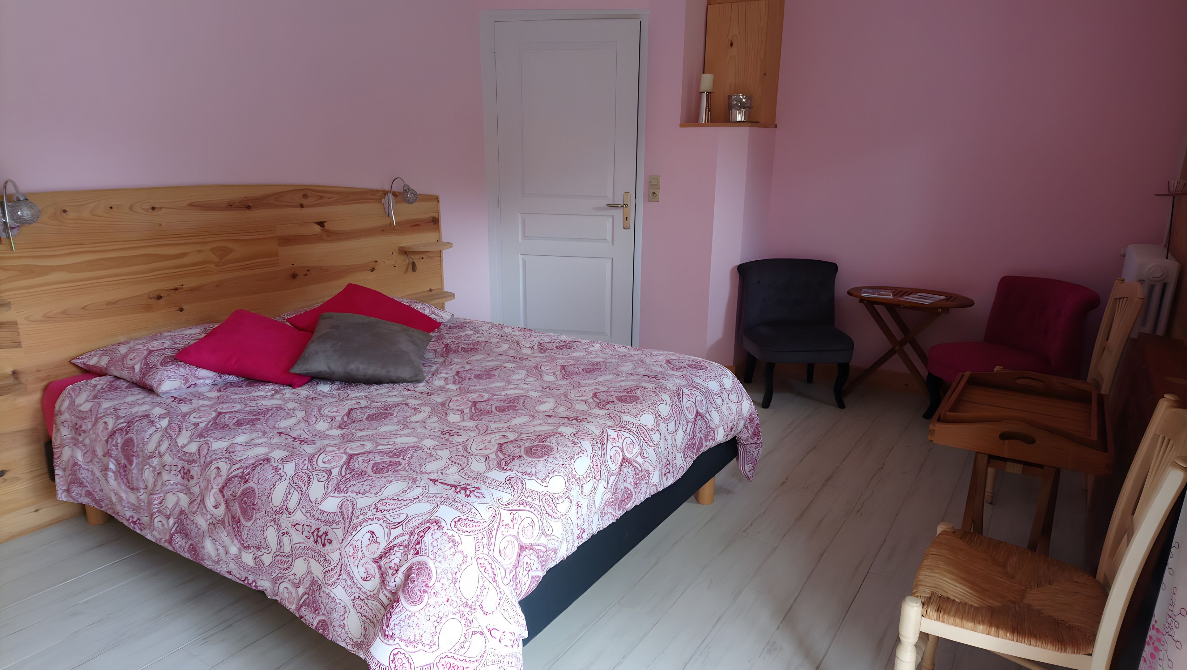 A bedroom with a wooden headboard, a bed with patterned bedding, a chair, a small table, and a door.