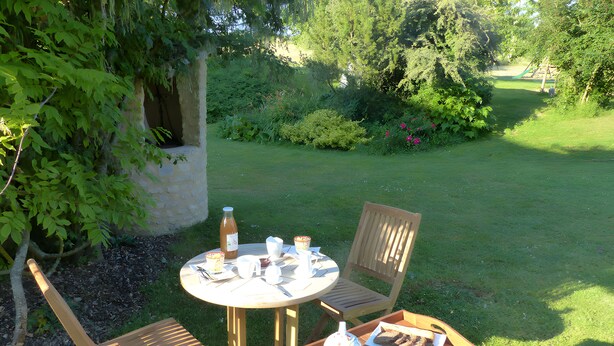 A round wooden table with chairs, a bottle, cups, and a plate with food, set in a grassy outdoor area with trees and a garden in the background. - Bed and Breakfast 'Chambre D'hôtes Bois De Rose' with Private Garden and Wi-Fi (La Hoguette)