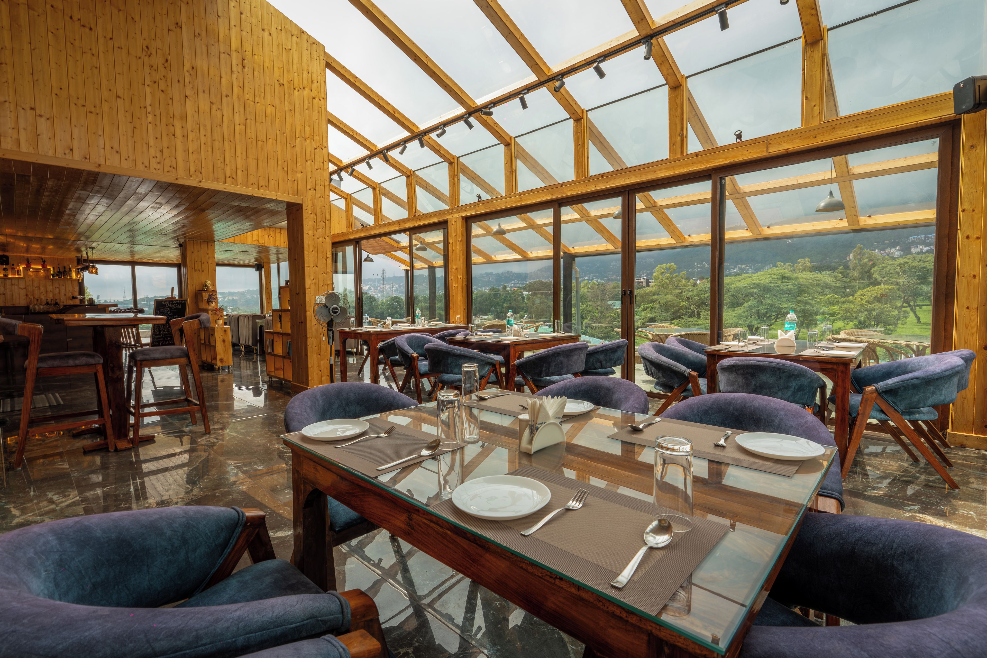 Un restaurante con techos altos y amplias ventanas que ofrecen vistas al campo. Las mesas están dispuestas con platos, cubiertos y copas.