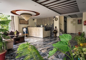 Un vestíbulo de hotel con una recepción, una araña de cristal, un sofá y diversas plantas.
