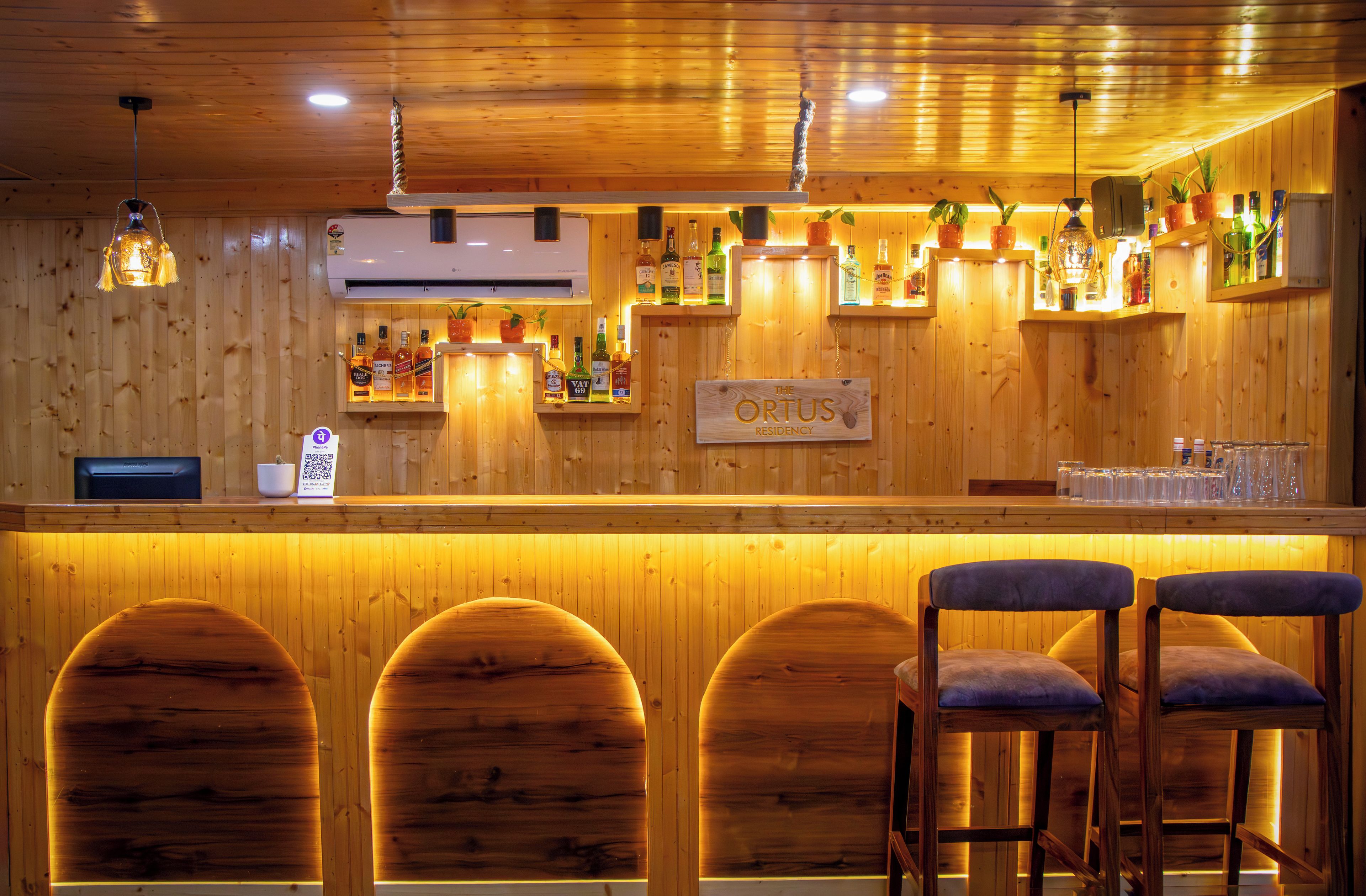 A bar area with wooden walls, a sign reading 'The Ortus Residency,' and multiple liquor bottles displayed on shelves.