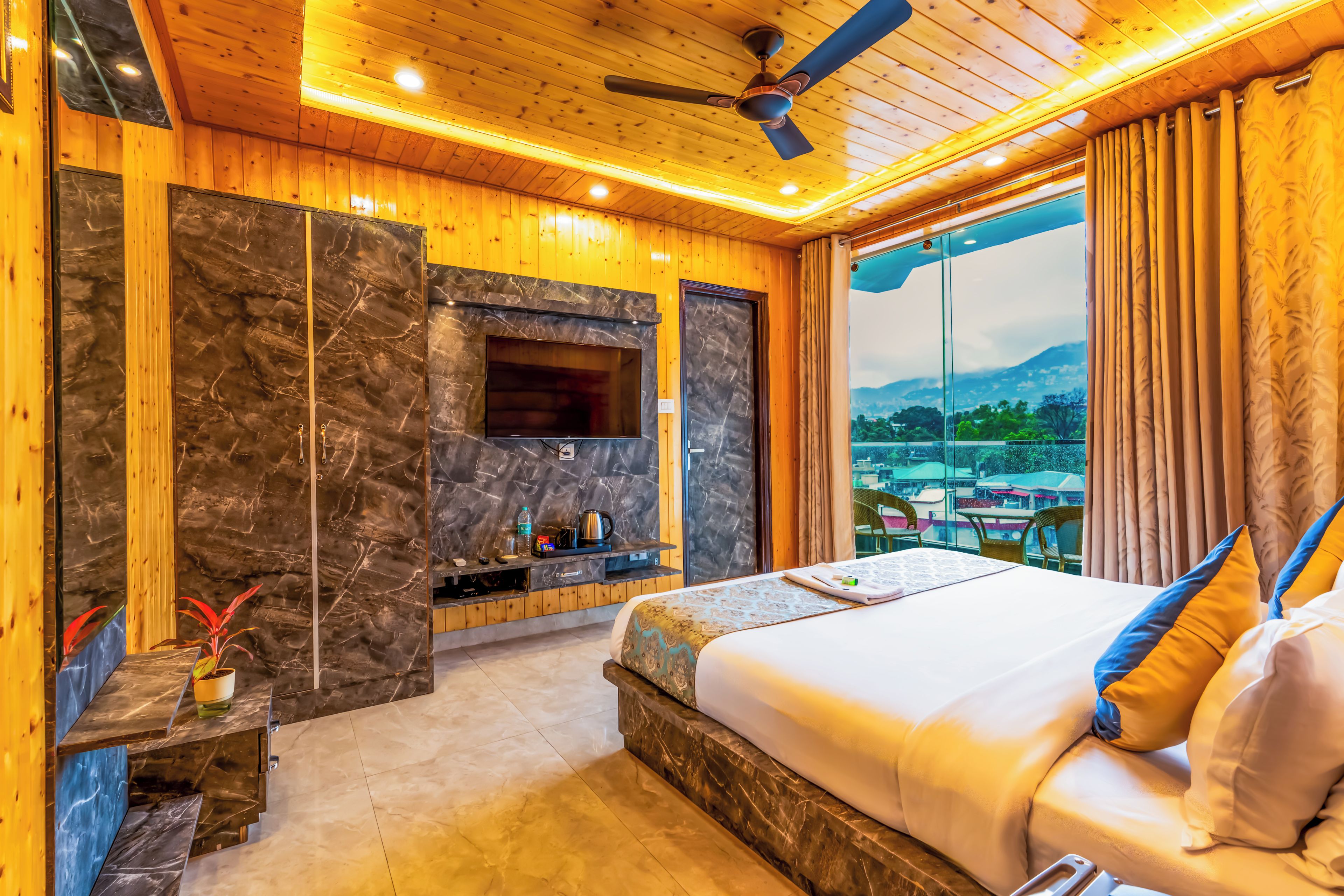 A hotel room with a large bed, a flat-screen TV mounted on a marble wall, a ceiling fan, and a view of a mountainous landscape.