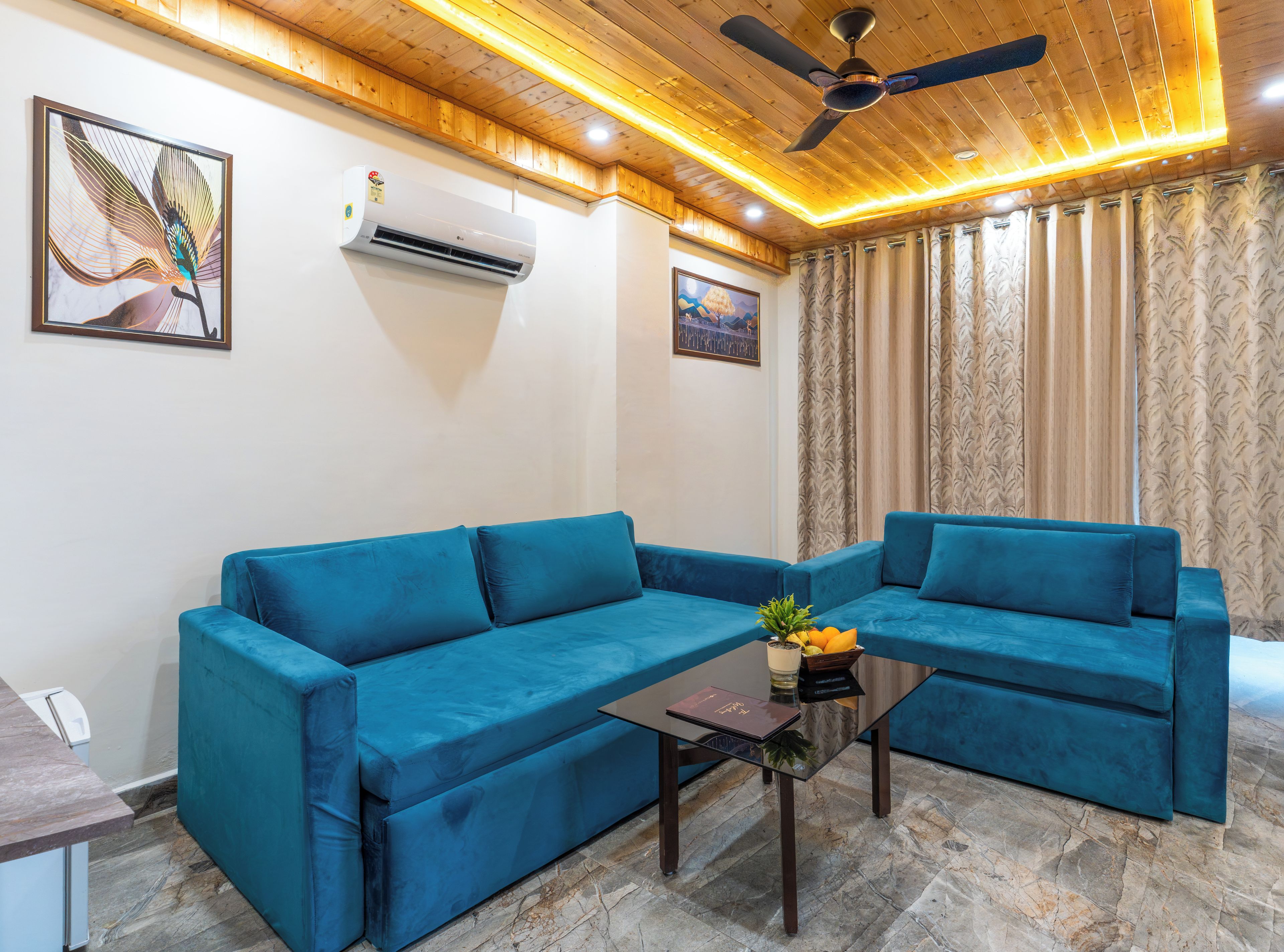 Ein Wohnzimmer mit einem blauen Ecksofa, einem kleinen Tisch mit Pflanze und Obst, und einem Deckenventilator.