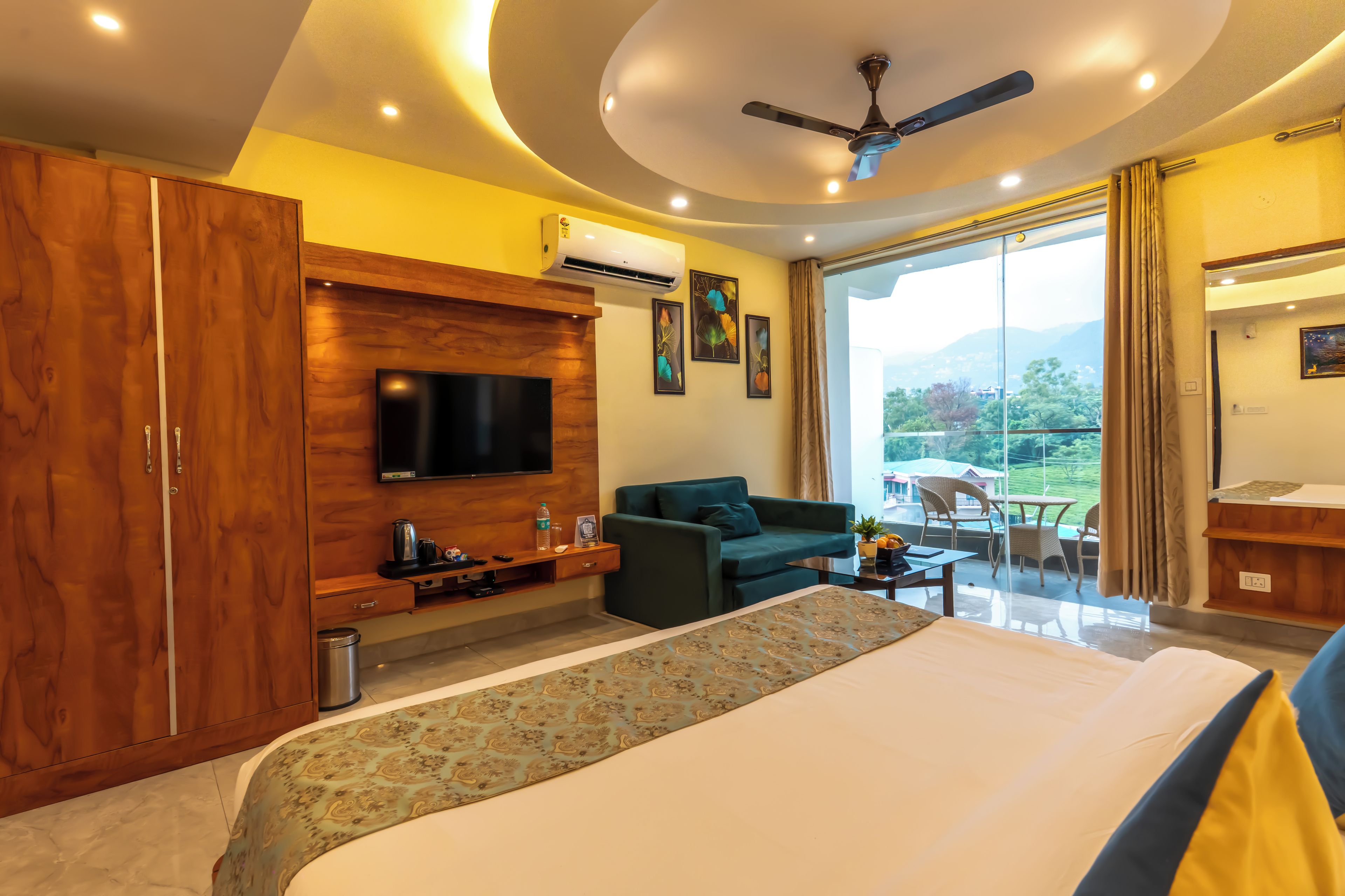 A hotel room with a large bed, a TV mounted on a wooden panel, a sofa, a small table, and a balcony with a view.