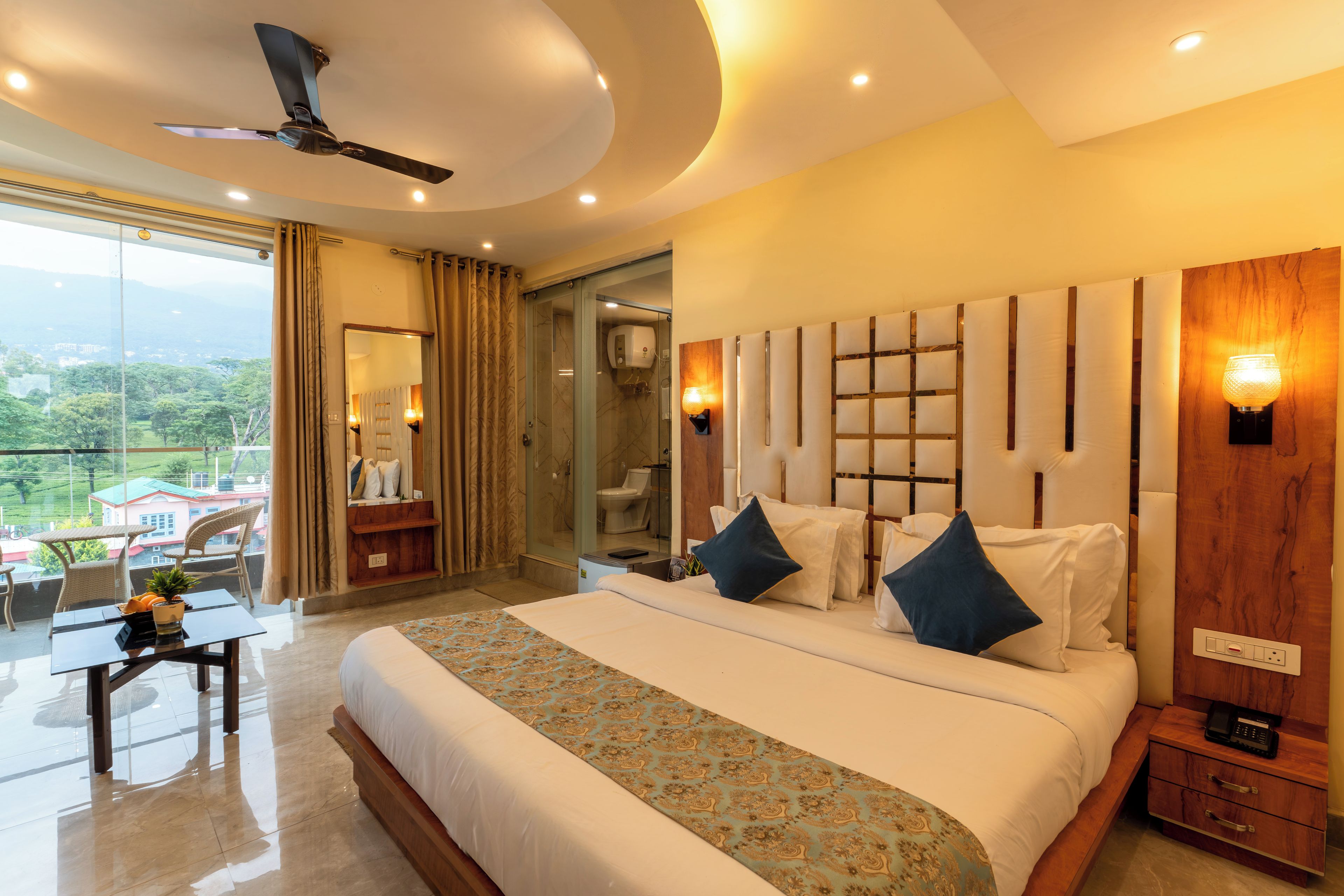 Une chambre d’hôtel avec un grand lit, une table de chevet, un bureau avec une chaise, une salle de bain avec une porte en verre et une vue sur l’extérieur.