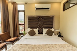 Habitación de hotel con cama, una silla, unidad de aire acondicionado, mesita de noche y ventana con cortinas.