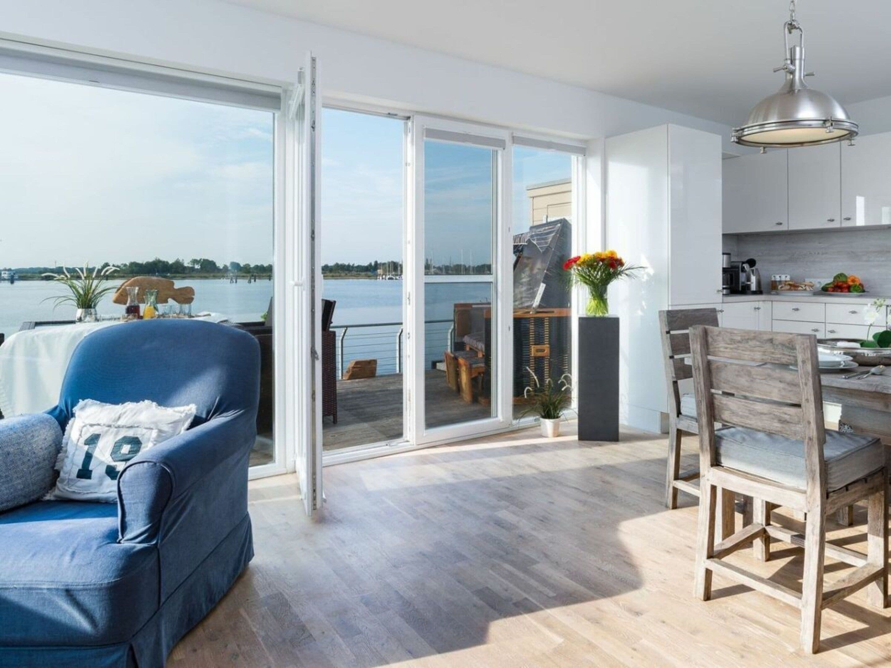 Ein modernes Wohnzimmer mit einer Couch, Esstisch und Stühlen, einer Küchenzeile und einer Glasschiebetür, die auf eine Terrasse mit Blick auf das Wasser führt.