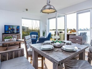 Ein Essbereich mit einem Holztisch, gedeckt für zwei Personen, einem Fernseher auf einem Holzständer und Blick auf das Wasser durch große Fenster.