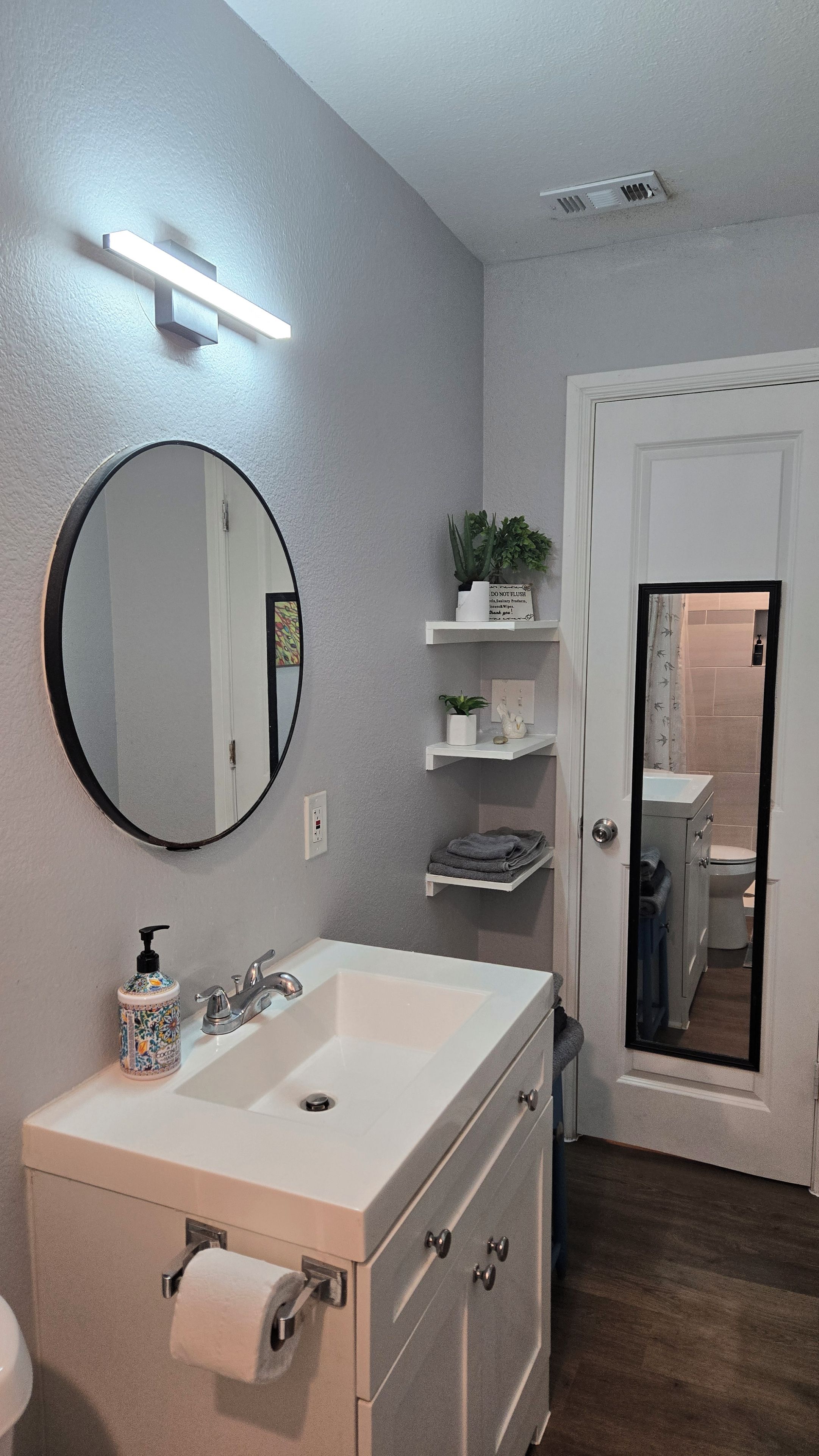 A bathroom with a white sink, a round mirror, a towel rack, and a door leading to a shower.