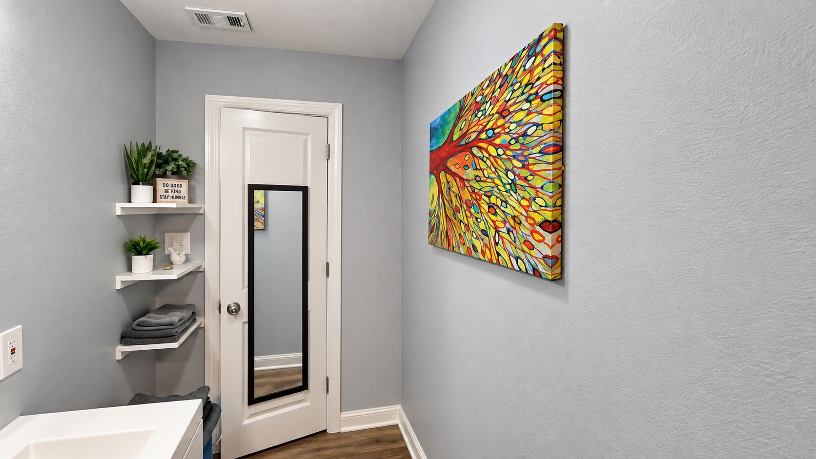 A bathroom with a white door, a mirror, a shelf with plants, and a colorful abstract painting on the wall.