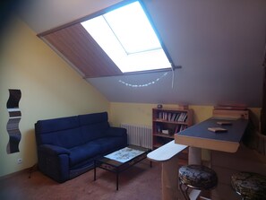 Une pièce chaleureuse avec un plafond en pente, un canapé bleu, une petite table, une bibliothèque et un radiateur.