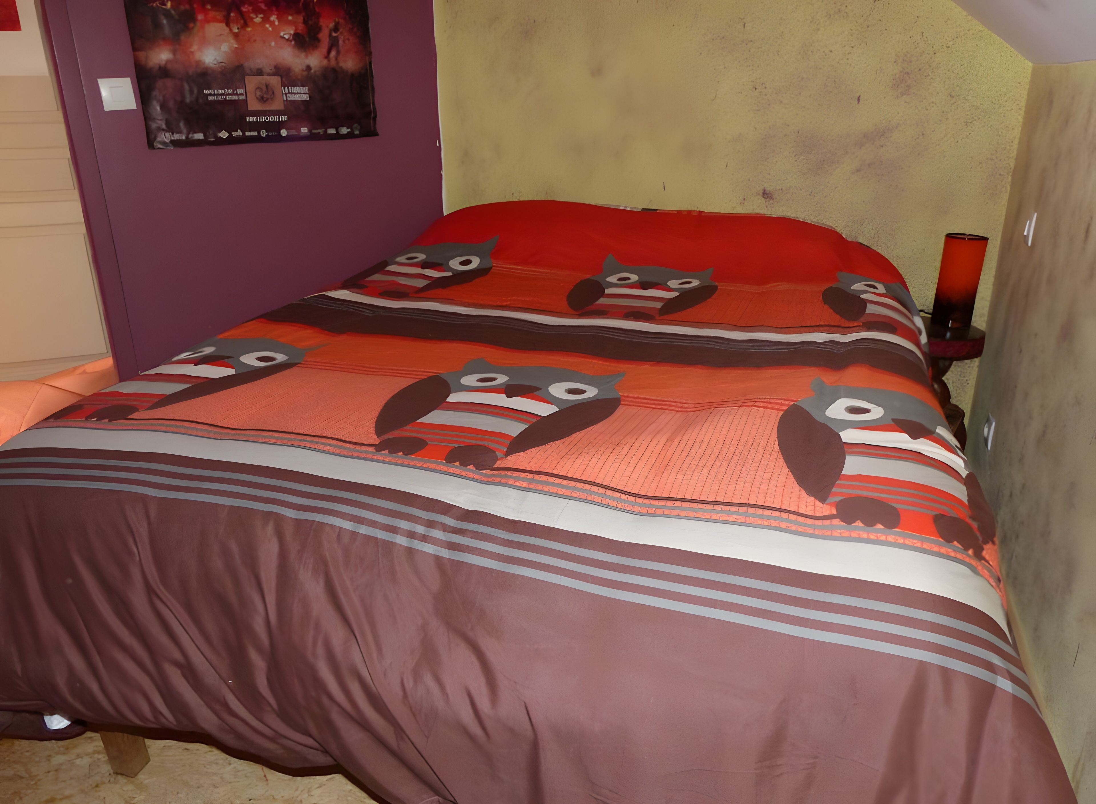 A bed with a patterned bedspread and pillows, a poster on the wall, and a bedside lamp.