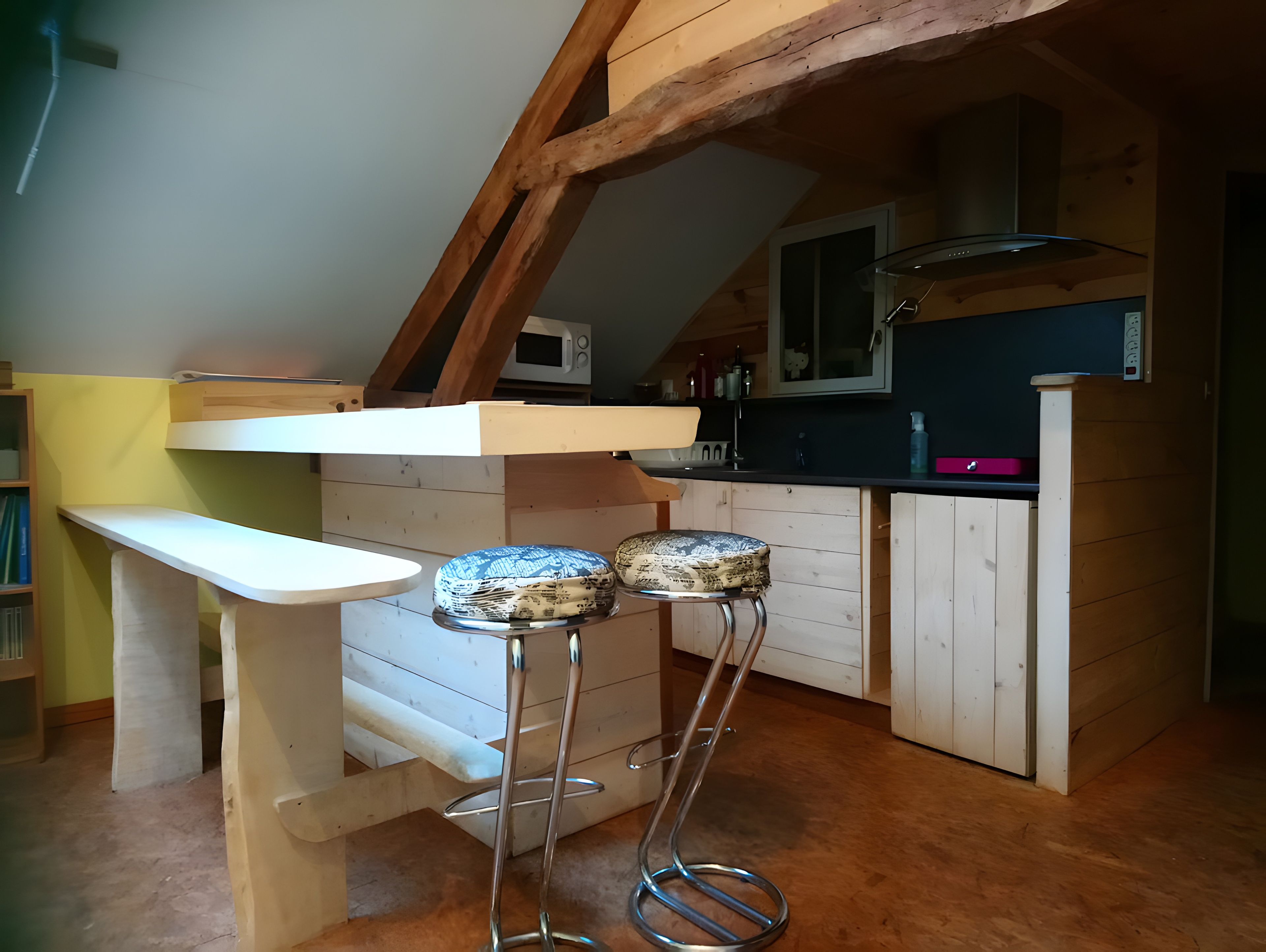 Cocina compacta con techo de madera, encimera blanca y microondas. Hay dos taburetes con cojines estampados.