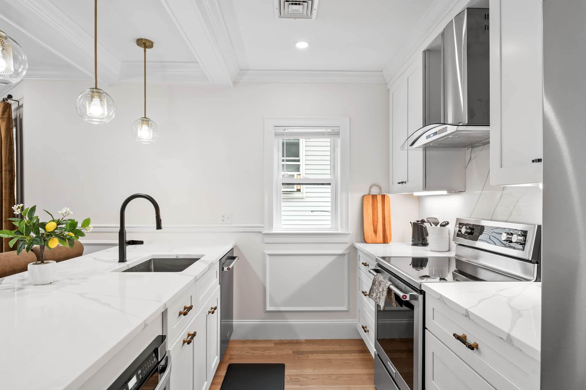 Une cuisine moderne avec des meubles blancs, un four encastré, une hotte de cuisson en acier inoxydable et des luminaires suspendus.