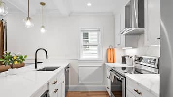 Une cuisine moderne avec des meubles blancs, un four encastré, une hotte de cuisson en acier inoxydable et des luminaires suspendus.