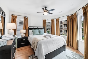 A hotel room with a bed, bedside tables, a lamp, a ceiling fan, and a window with curtains. - Designer Condo in Jamaica Plain | Steps to Forest Hills (Boston)