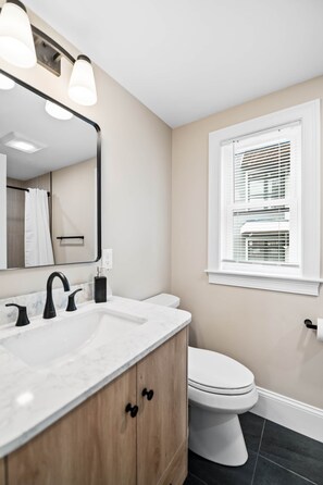 A modern bathroom with a white sink, black faucet, and wooden cabinet. There is a toilet and a window with blinds. - Designer Condo in Jamaica Plain | Steps to Forest Hills (Boston)