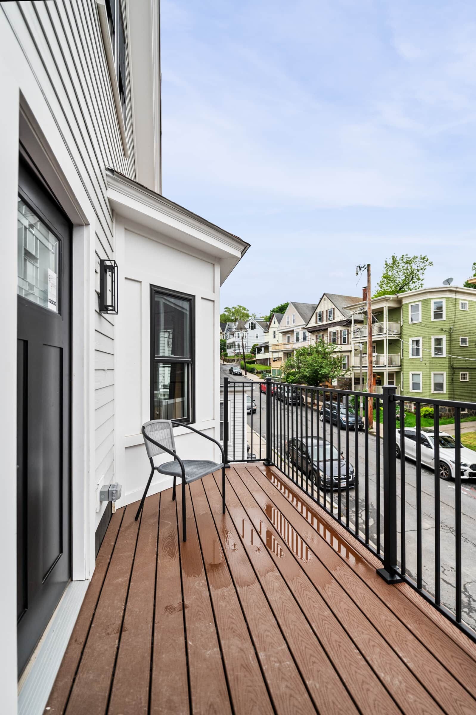 A wooden deck with a chair, a black metal railing, and a view of a street with houses.
