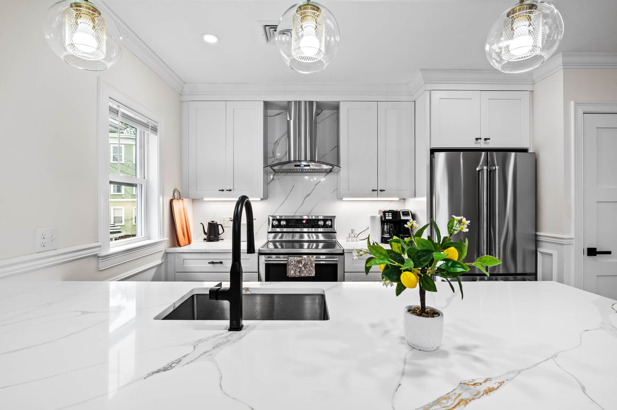 A modern kitchen with white cabinetry, stainless steel appliances, a marble countertop, and pendant lighting.