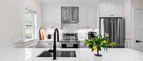Una cocina moderna con muebles blancos, electrodomésticos de acero inoxidable, encimera de mármol e iluminación colgante.