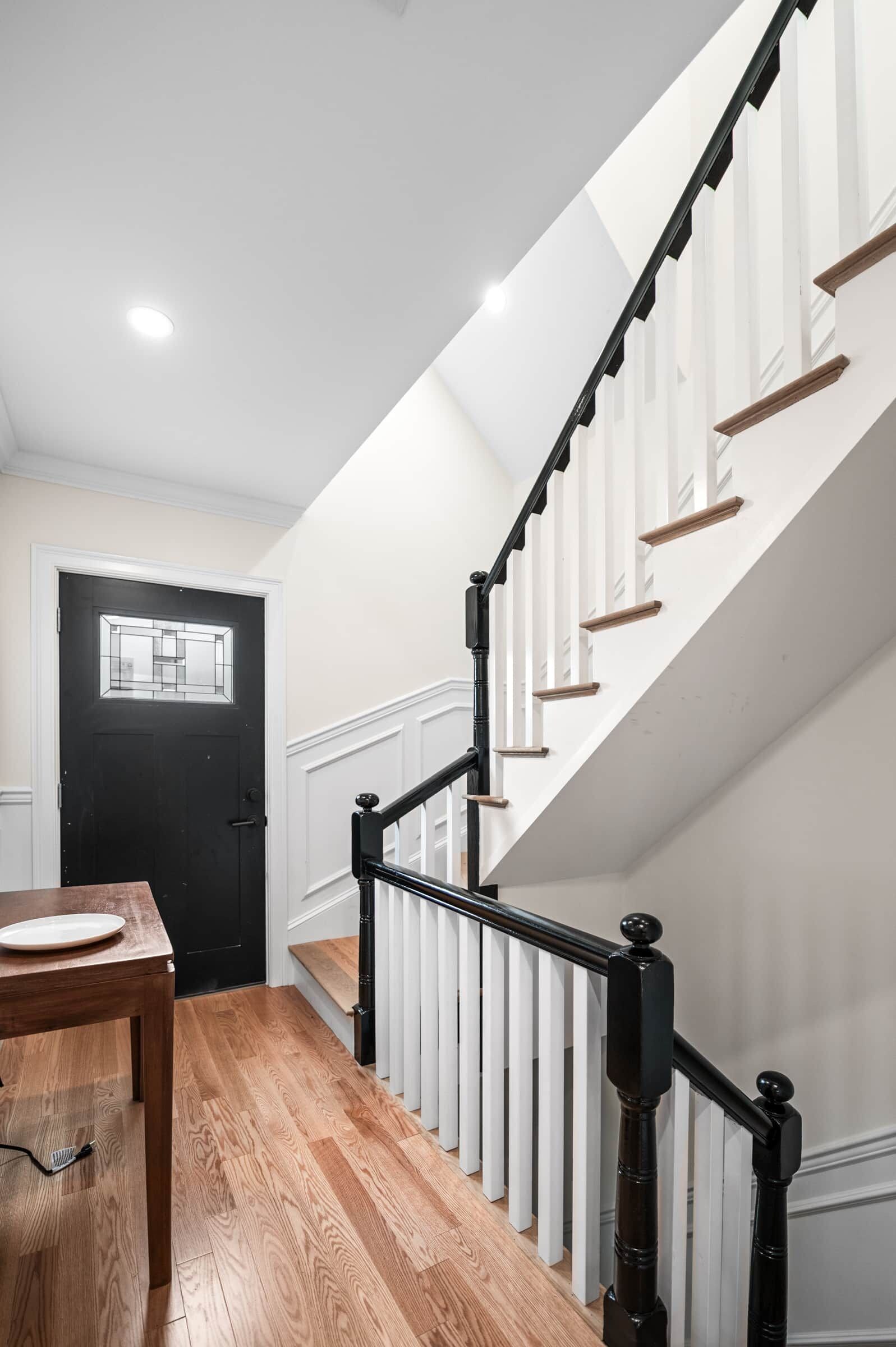A modern interior with a staircase, wooden flooring, a dining table, and a black door.
