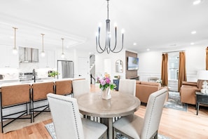 A modern kitchen with stainless steel appliances, a dining area with a round table and chairs, and a living room with a sofa and a TV. - Designer Condo in Jamaica Plain | Steps to Forest Hills (Boston)