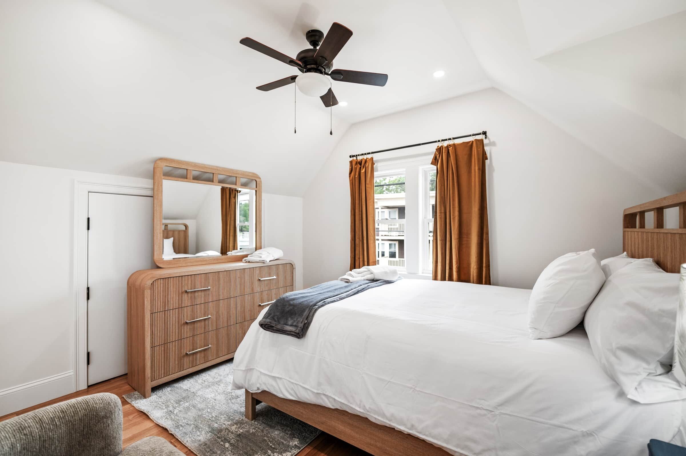 Une chambre à coucher avec un lit, une commode en bois, un ventilateur de plafond et une fenêtre avec des rideaux.