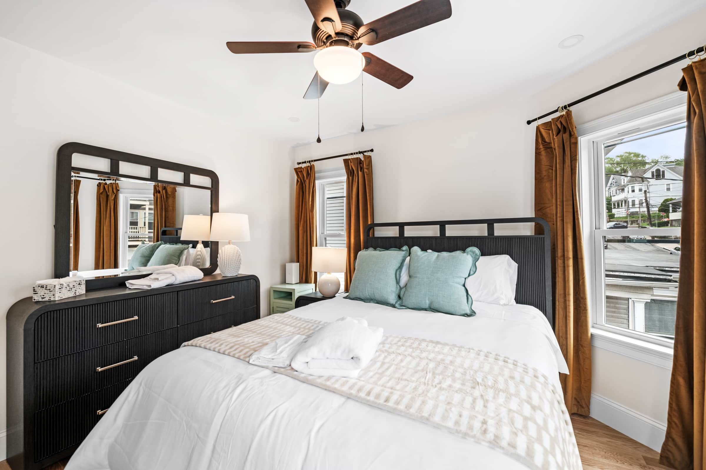 Une chambre d’hôtel avec un lit, une commode, un miroir, un ventilateur de plafond et une fenêtre avec des rideaux.