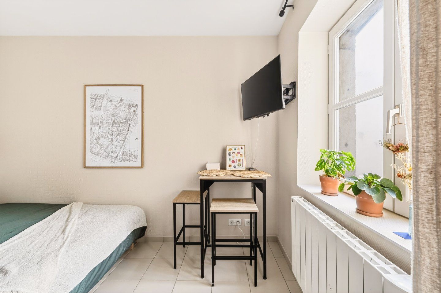 A small, well-lit room with a bed, a desk with a chair, a flat-screen TV mounted on the wall, and a window with potted plants.