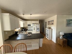 Una cocina compacta con armarios blancos, una isla central y una zona de comedor con sillas de madera.