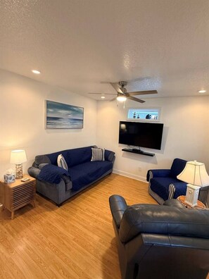 A living room with a blue sofa, a black leather chair, a flat-screen TV, a ceiling fan, and a wooden floor.