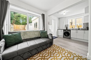 A modern apartment with a grey sofa, a kitchen with a washing machine, and a view of a garden. - Spacious 5 Bedrooms 2.5 Bathrooms | 8 Beds | WiFi | Driveway | Atherton M46 (Atherton)