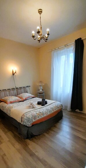 A bedroom with a bed, a nightstand, a lamp, a chandelier, and a window with curtains. - La Forge- Gite 4 pers- face au canal du Berry- Wifi (Thénioux)