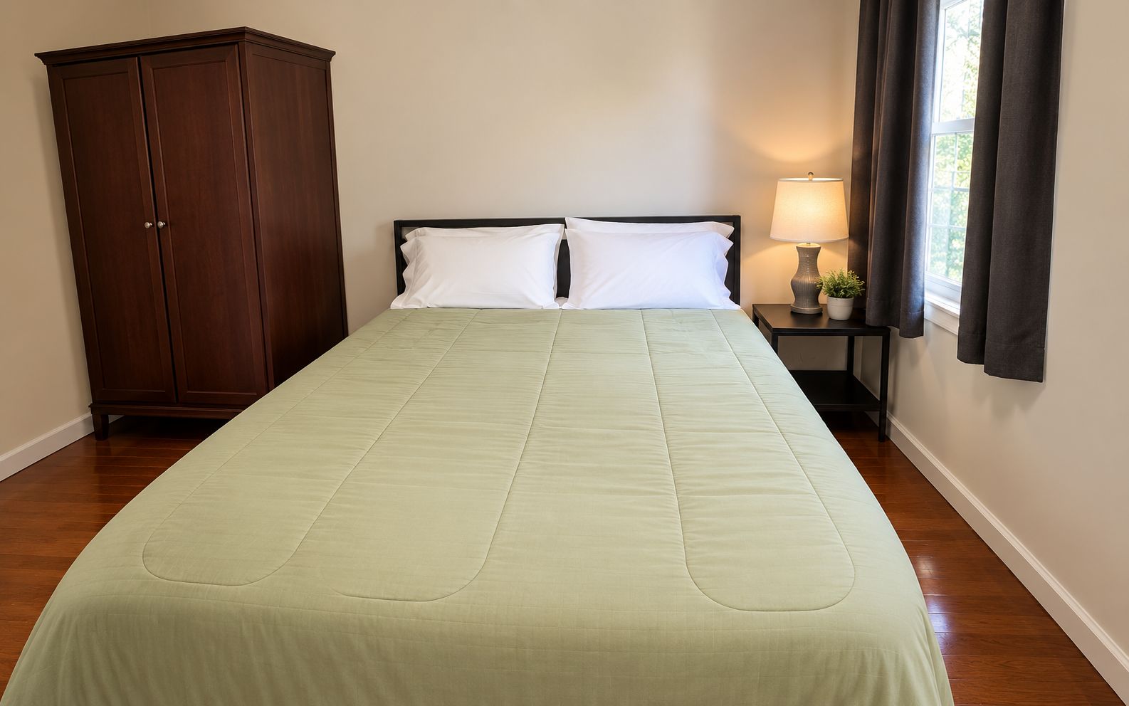 A single bed with a green bedspread and two white pillows, a wooden wardrobe, a bedside table with a lamp, and a window with curtains.