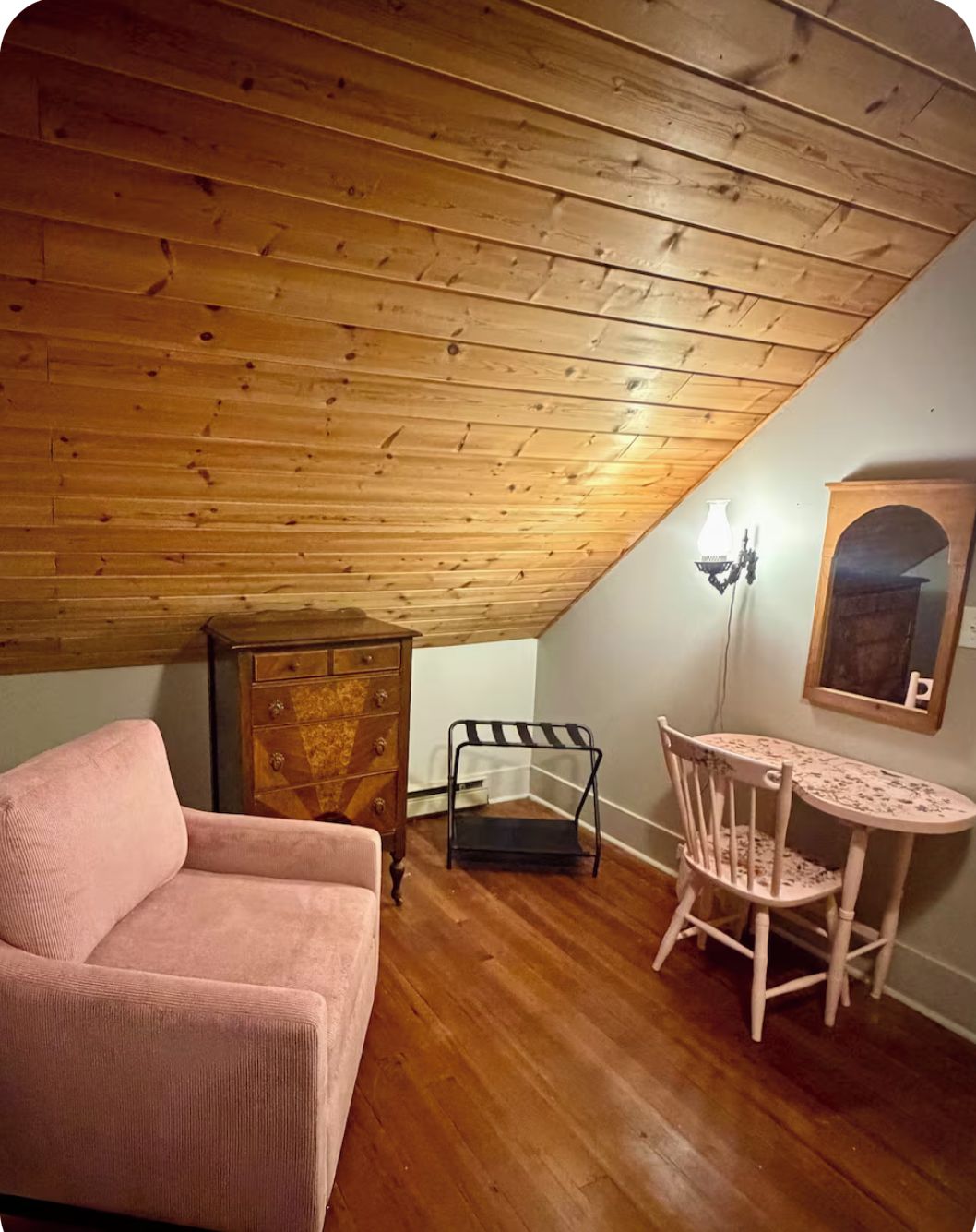 Ett mysigt rum med ett sluttande tak, en byrå i trä, en rosa soffa, ett litet bord med en stol och en spegel som hänger på väggen.