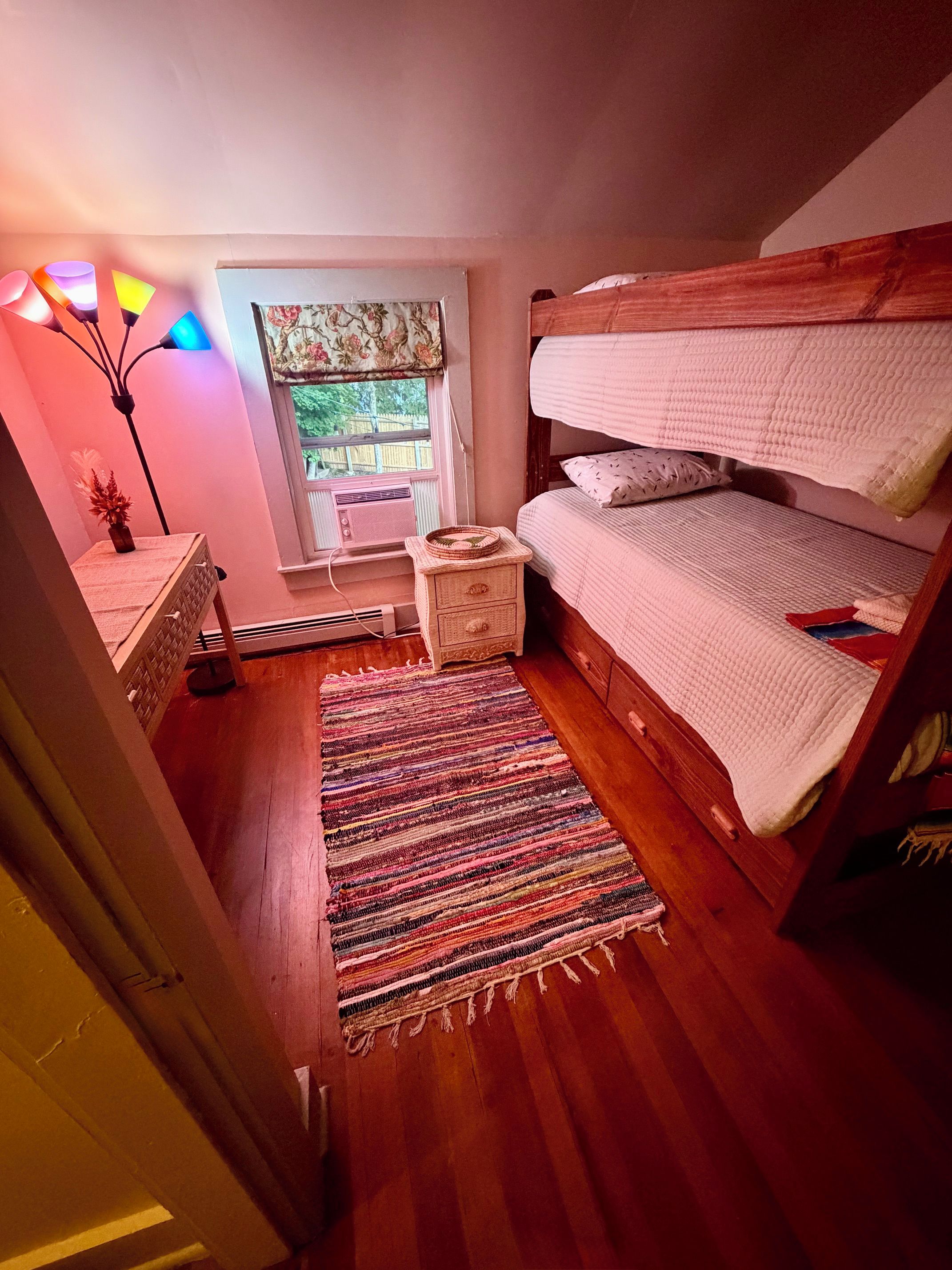A bedroom with a bunk bed, a small bedside table, a lamp, a window with a view, and a colorful rug.