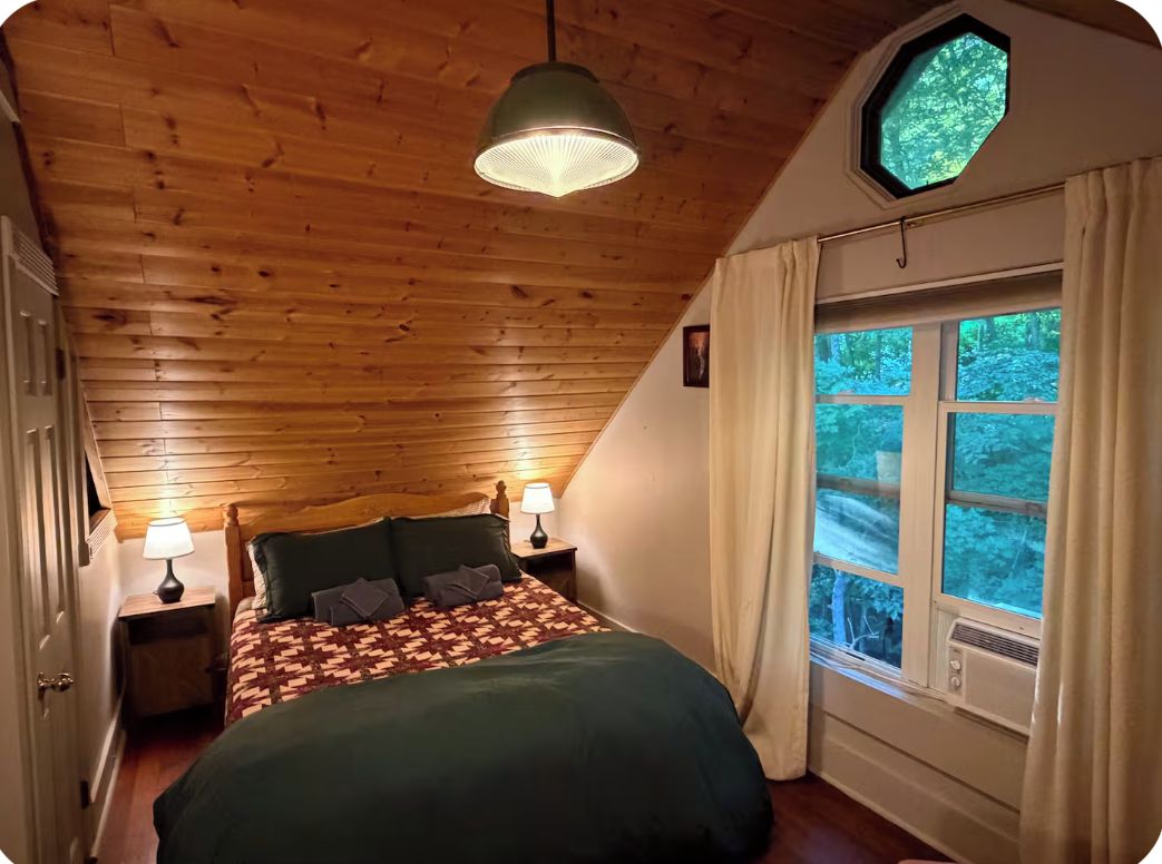 A bedroom with a wooden ceiling, a bed with a patterned blanket, two bedside lamps, and a window with curtains.