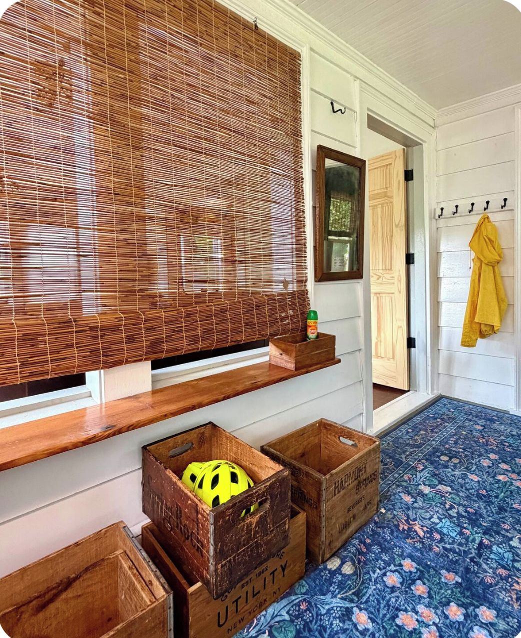 A room with a large window covered by bamboo blinds, wooden crates on the windowsill, a yellow towel hanging on a rack, and a floral-patterned rug on the floor.