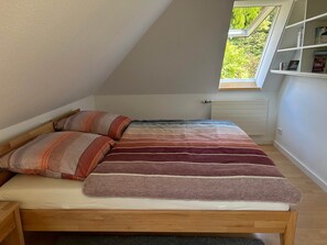 Ein Schlafzimmer mit schrägem Dach, einem Fenster, einem Bücherregal und einem Bett mit gestreifter Bettwäsche.