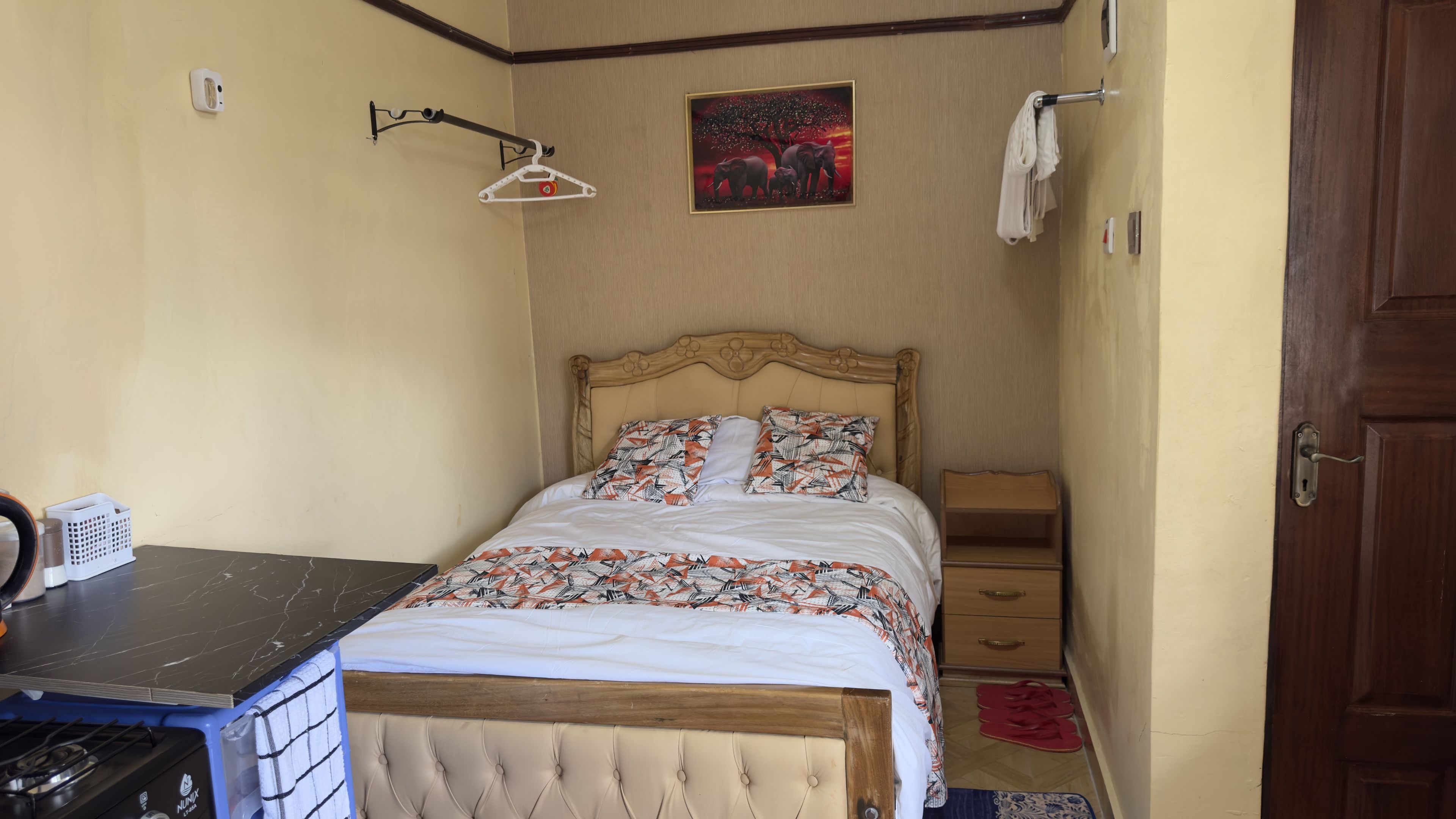 A bedroom with a bed, a nightstand, a wall-mounted towel rack, a painting, and a small wooden cabinet.