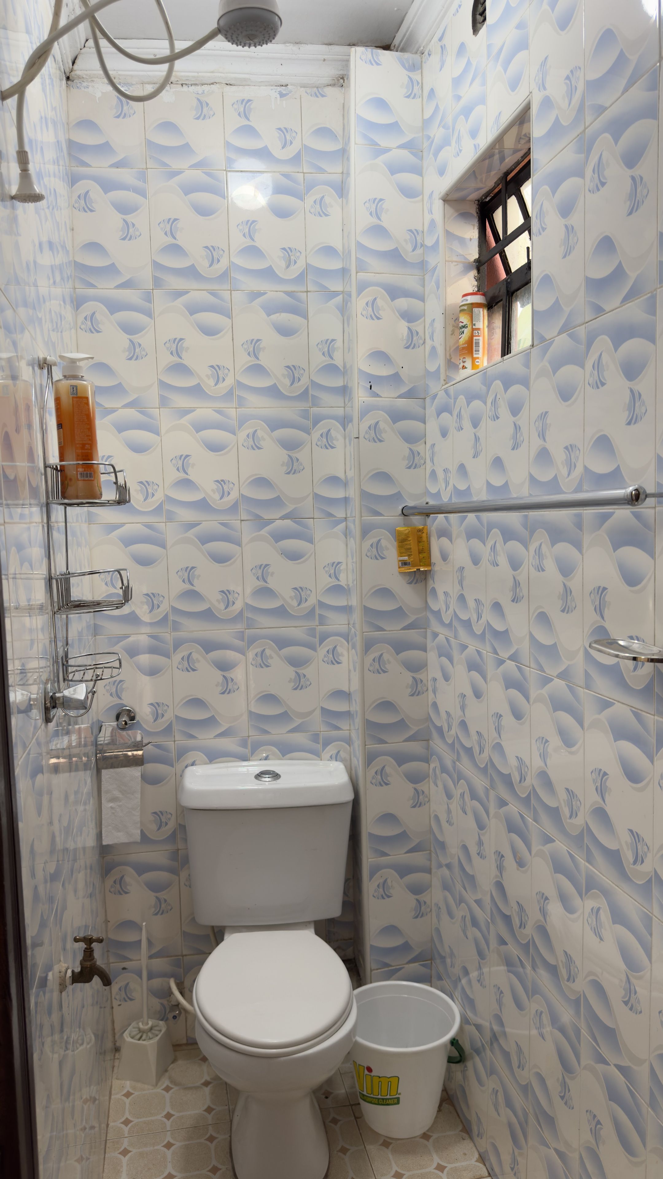 Une salle de bain avec des toilettes, une douche et un lavabo. Un flacon de savon liquide est accroché au mur et un seau blanc est posé sur le sol.