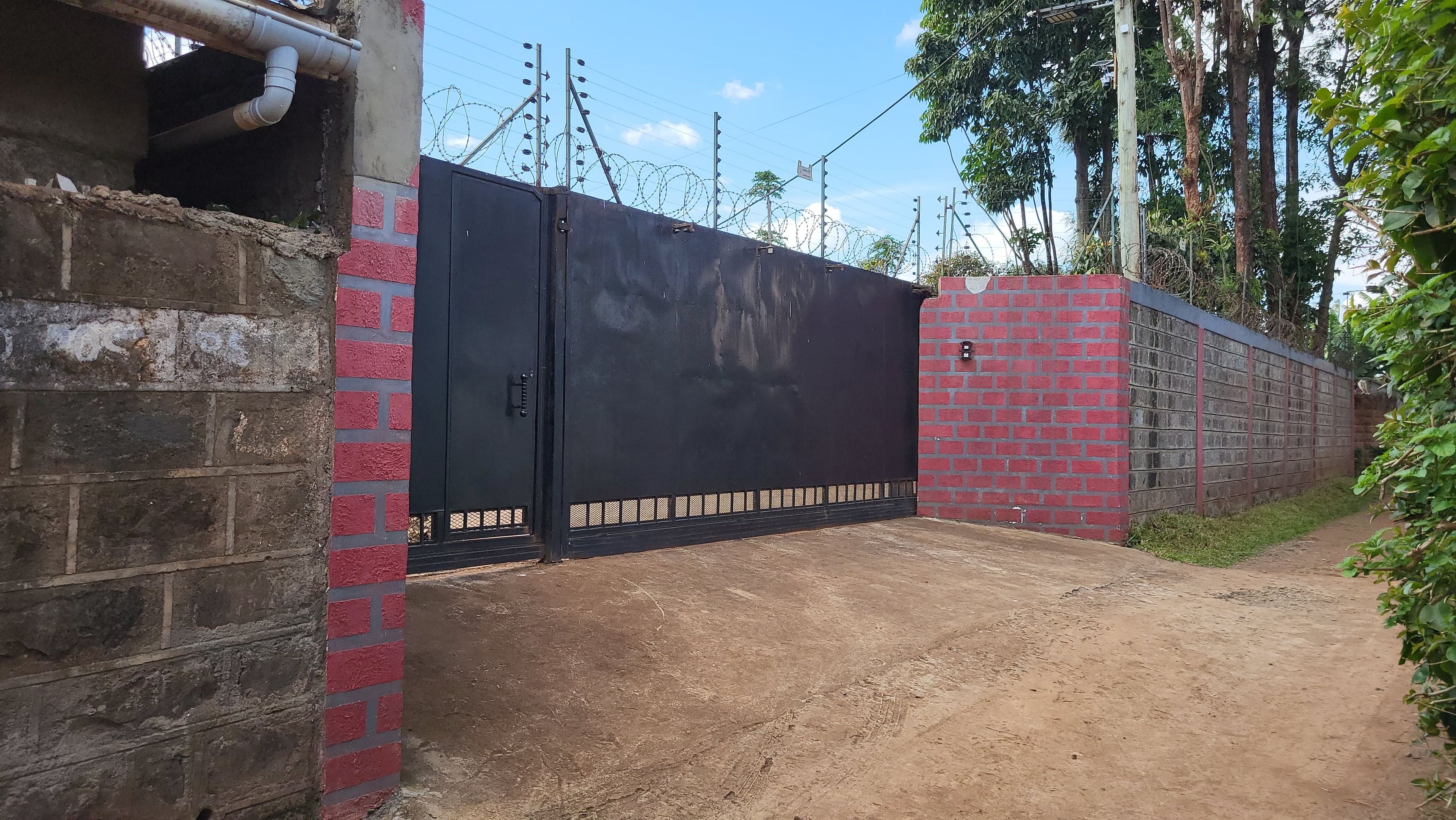 Una entrada protegida por un portón de metal negro, un muro de ladrillo y un camino de tierra que se adentra en un área boscosa.