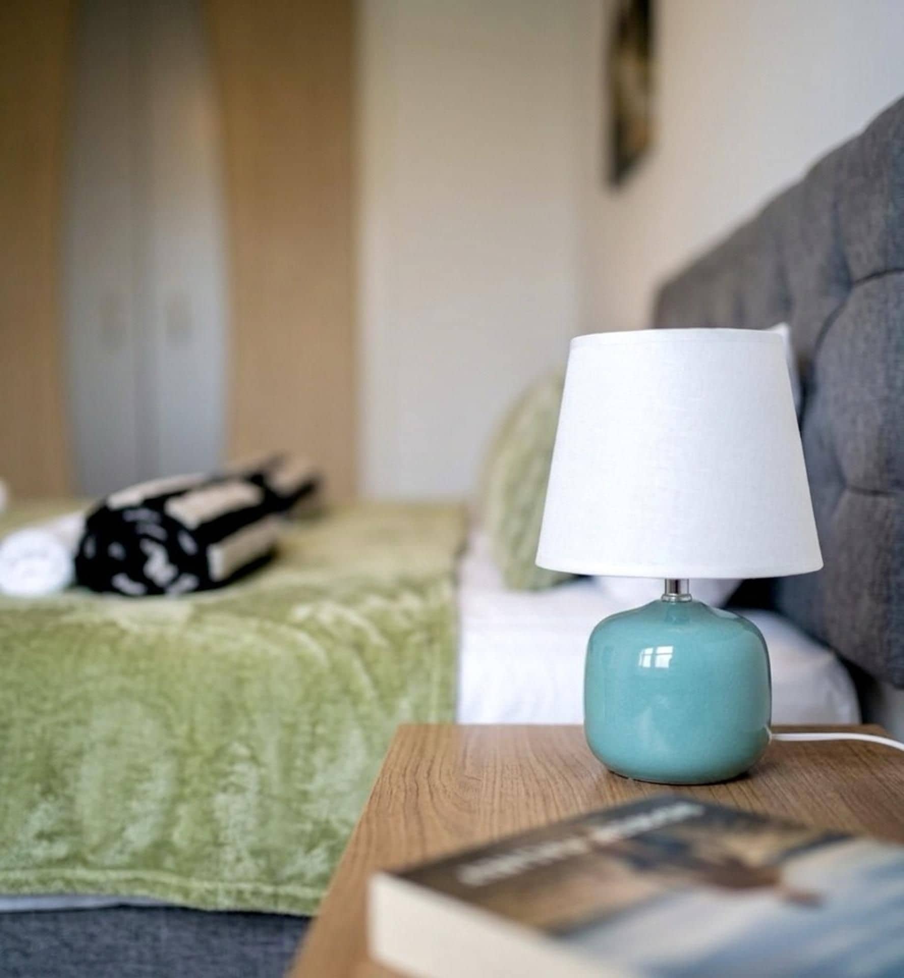 A neatly made bed with a green blanket, a grey headboard, and a white pillow. A teal lamp with a white shade is on a wooden nightstand beside the bed.