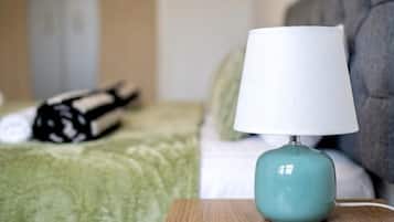 A neatly made bed with a green blanket, a grey headboard, and a white pillow. A teal lamp with a white shade is on a wooden nightstand beside the bed.