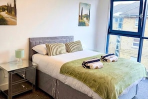A bedroom with a bed, pillows, a bedside table with a lamp, and a window.