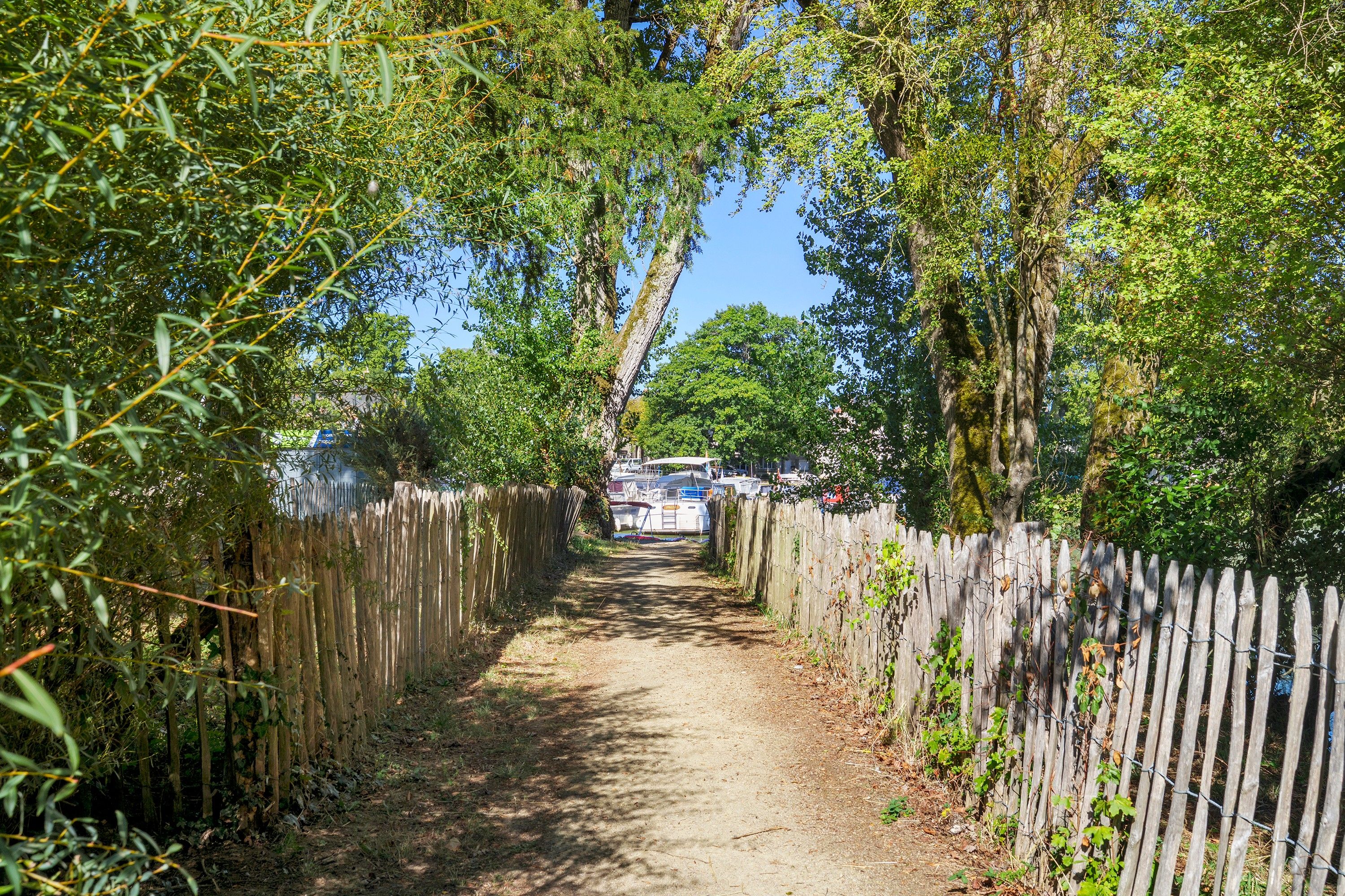 A narrow dirt path flanked by wooden fences, leading to a dock area with boats.
