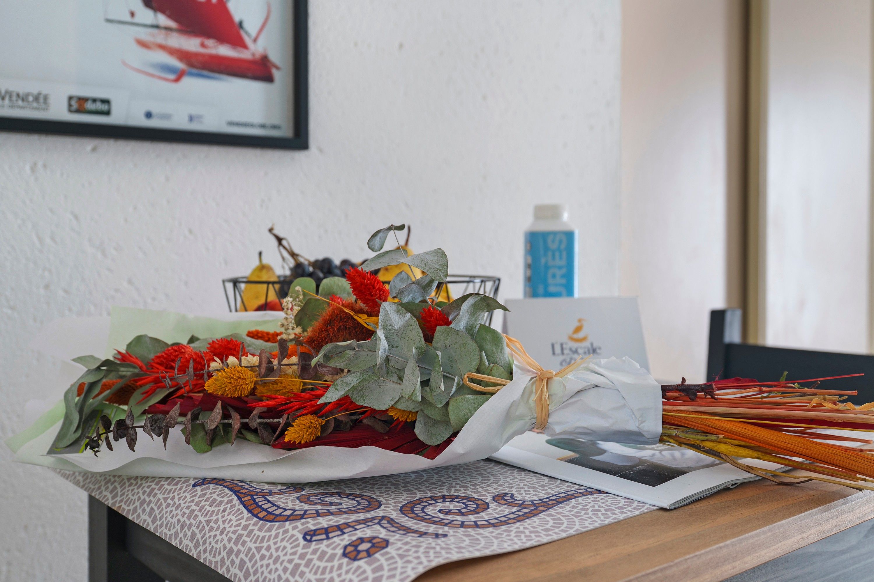 Una mesa con un arreglo floral, una botella y un cuadro enmarcado.
