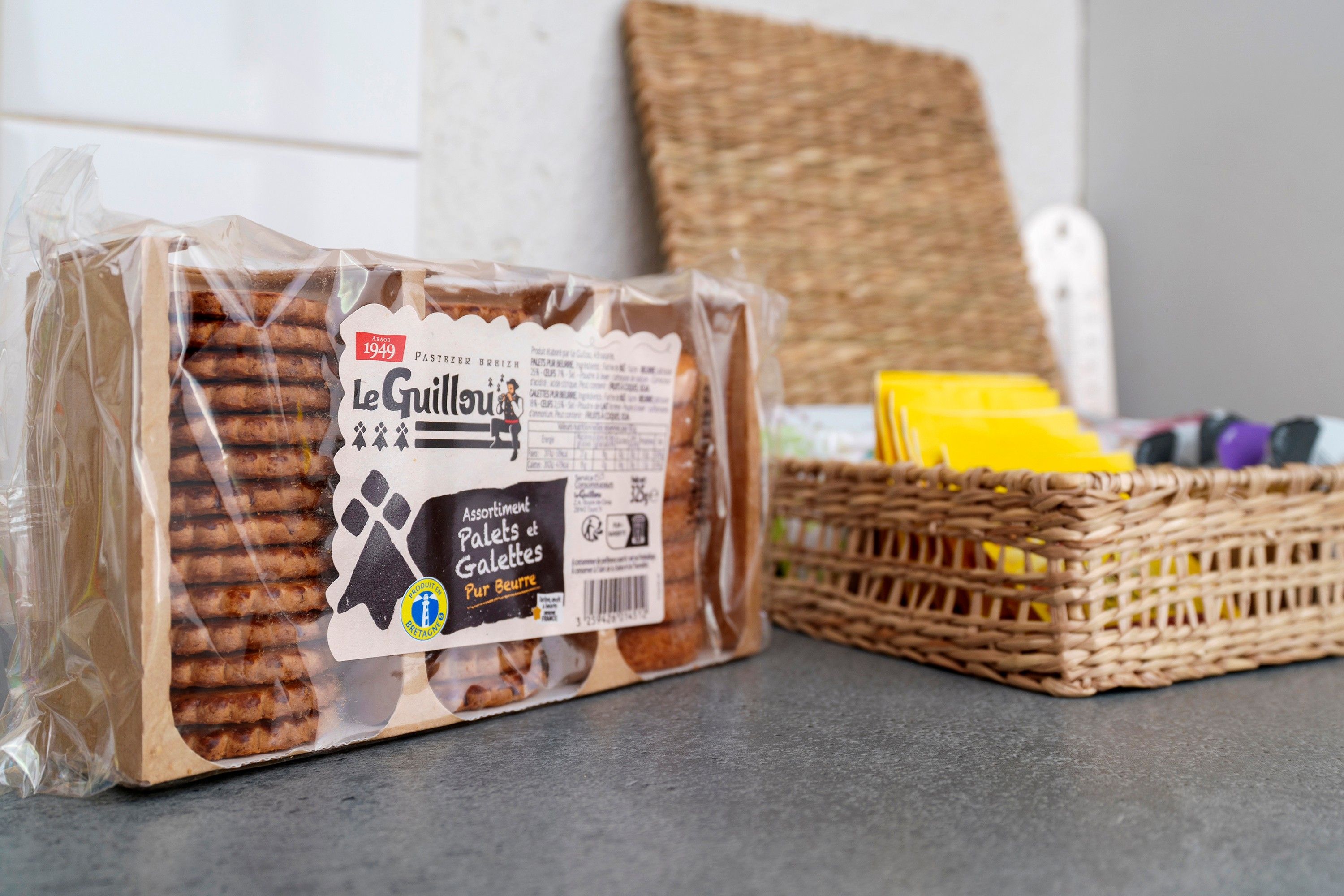 Eine Packung Le Guillon Kekse mit Etikett, platziert auf einem Tisch neben einem Korbwannen geflochtenen Korb mit gelb und violetten Gegenständen.